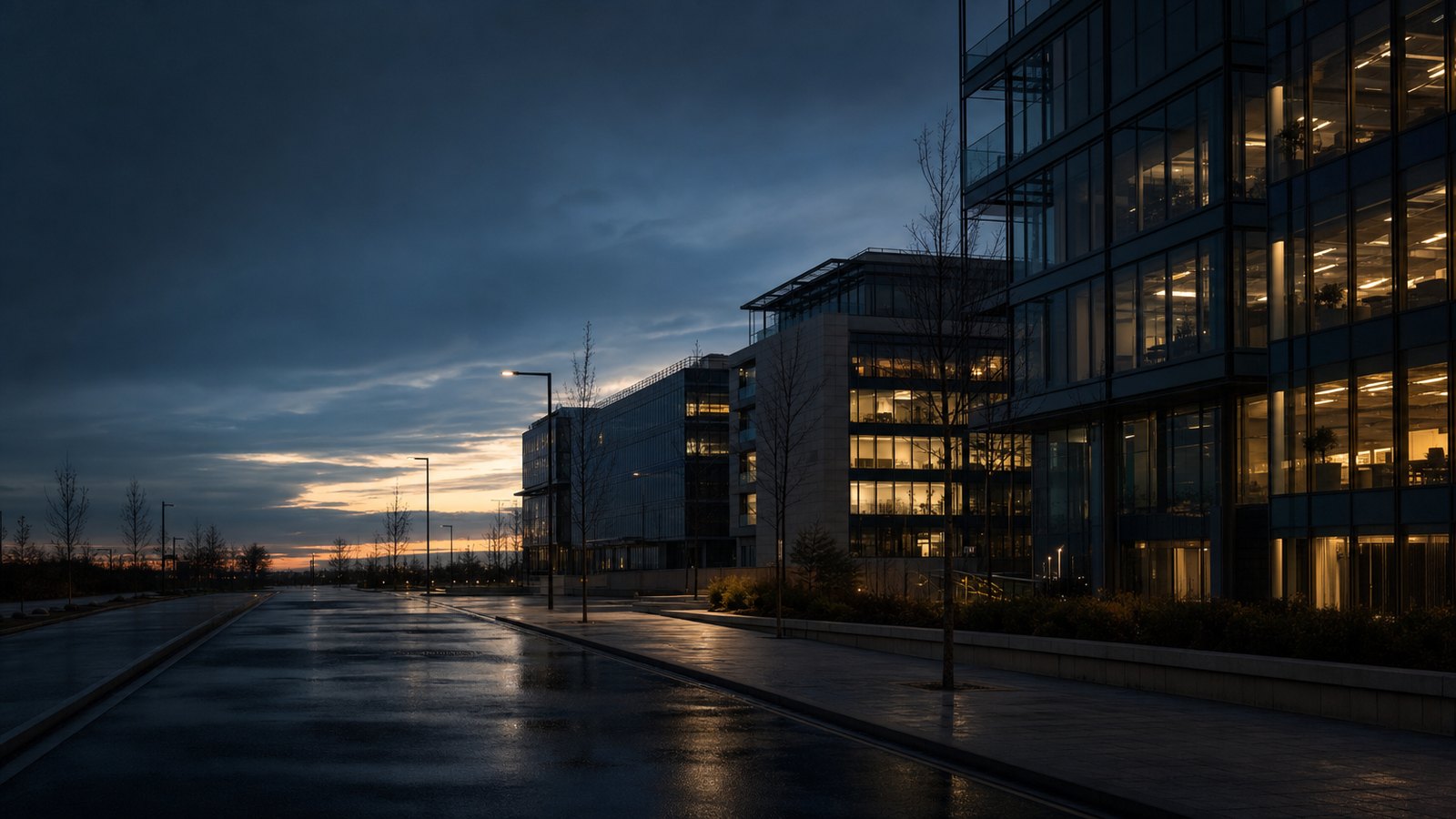 A modern south Dublin office quarter at dusk, lights on in scattered windows, wet road after rain.